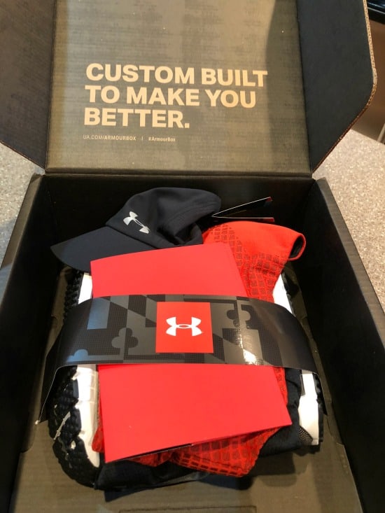 a black box with the lid open and a stack of red, black and white clothes strapped together with a paper that has the Under Armour logo